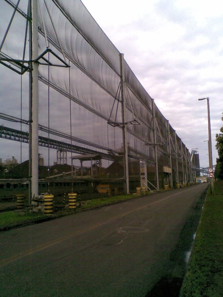 A long industrial wind fence stretches alongside a paved road with green grass on one side
