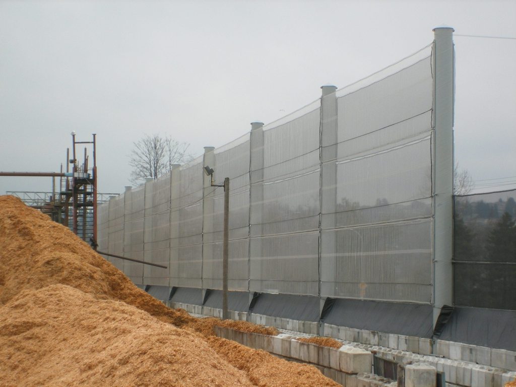 A tall, translucent wind fence stands next to a large pile of wood chips