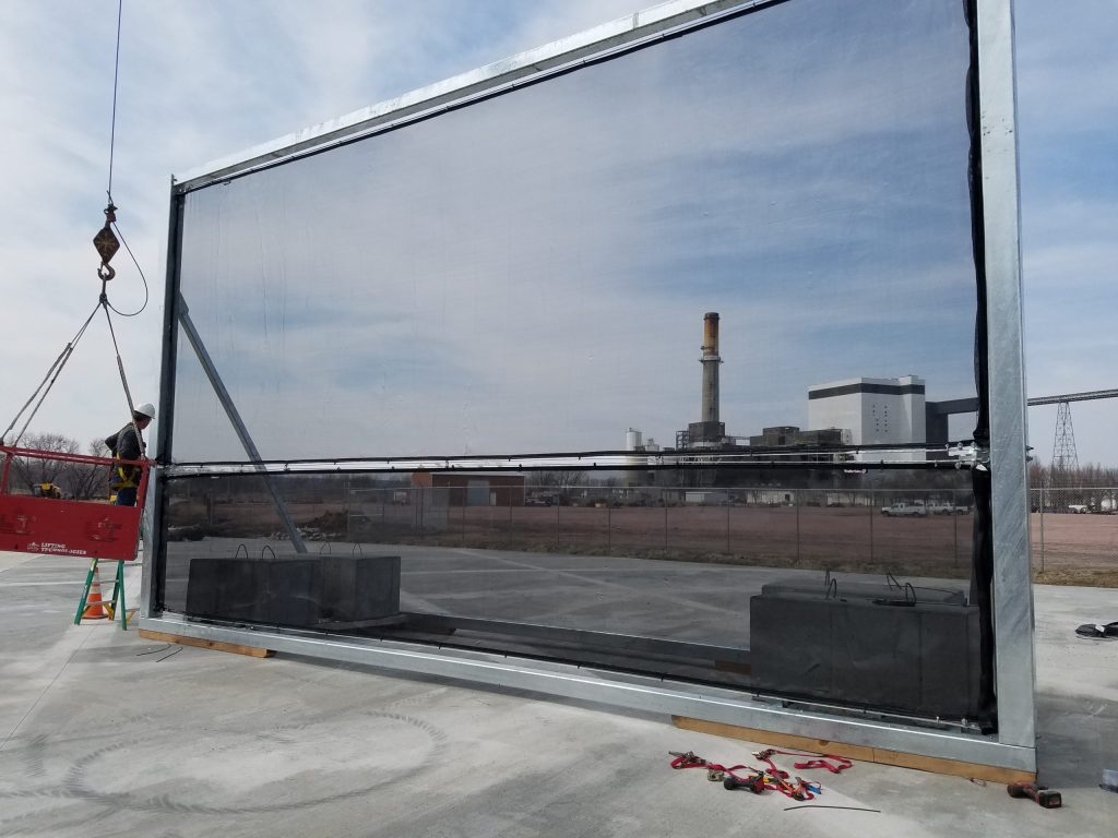 A large industrial wind fence being installed with a crane and worker in safety gear