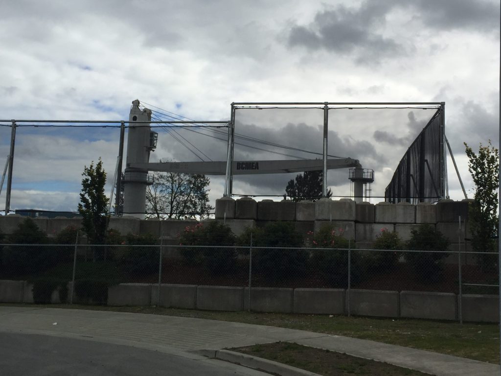 A large industrial crane stands behind a chain-link fence and concrete barriers