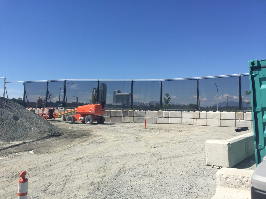 Construction site with orange machinery, gravel, concrete barriers, and a black mesh fence