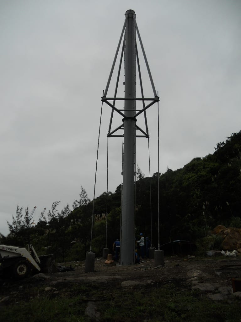 A tall metal pole with guy wires and a ladder, surrounded by construction equipment and workers