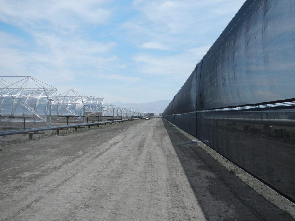 A long dirt road stretches between a row of metal structures and a tall black fence