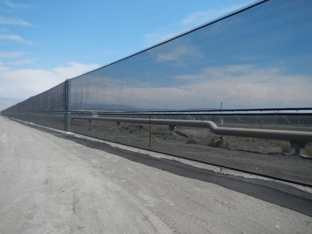 A long black mesh fence stretches alongside a dirt road under a clear sky