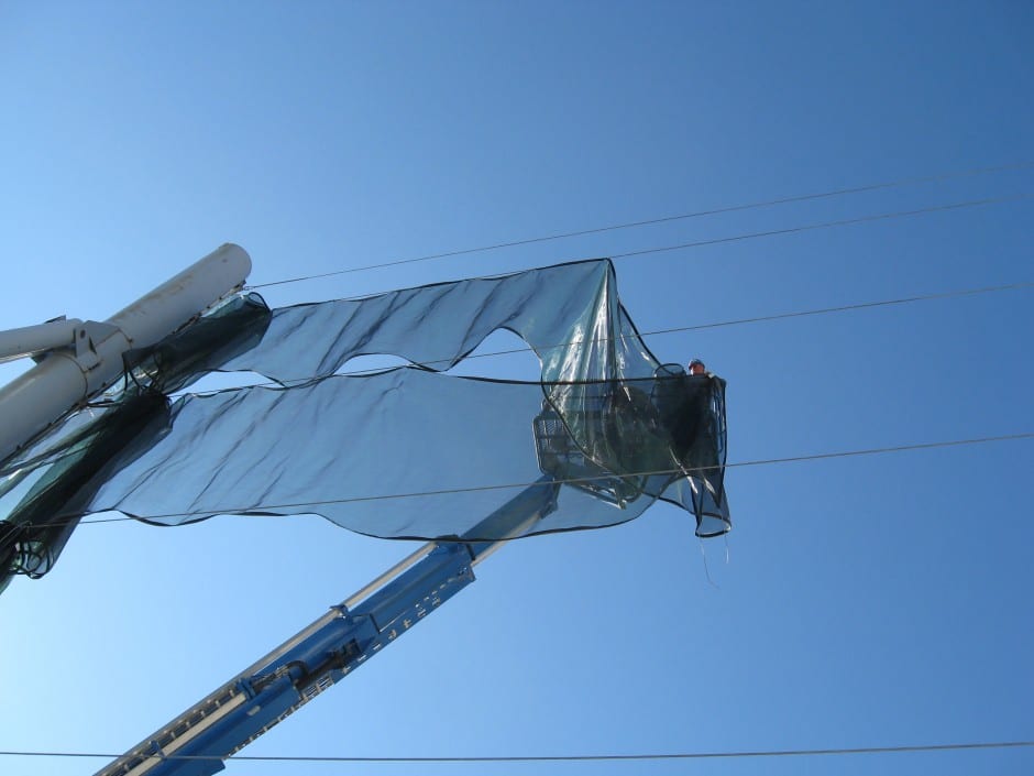 A person in a crane basket adjusts a translucent wind fence against a clear blue sky