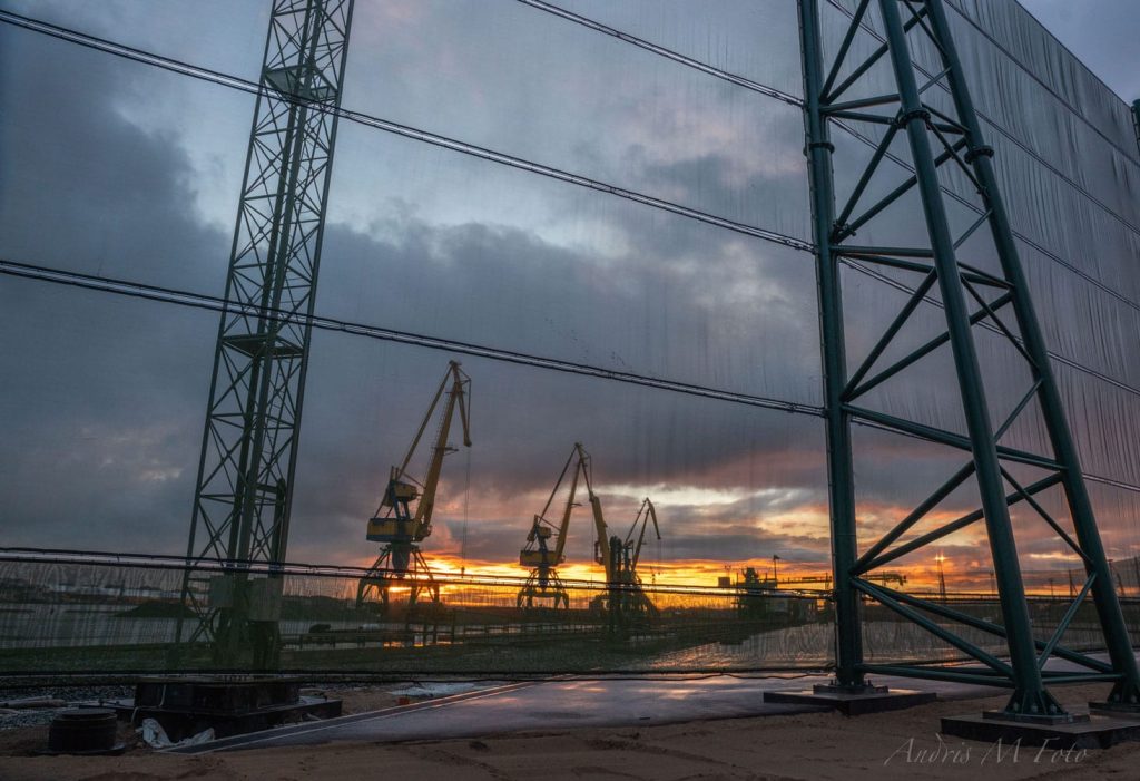 Cranes silhouetted against a sunset, viewed through a mesh fence with metal supports