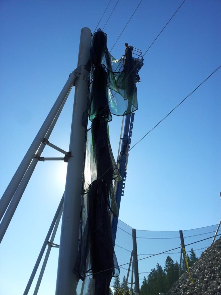 A person stands on a lift installing a green mesh netting on tall poles against a clear blue sky
