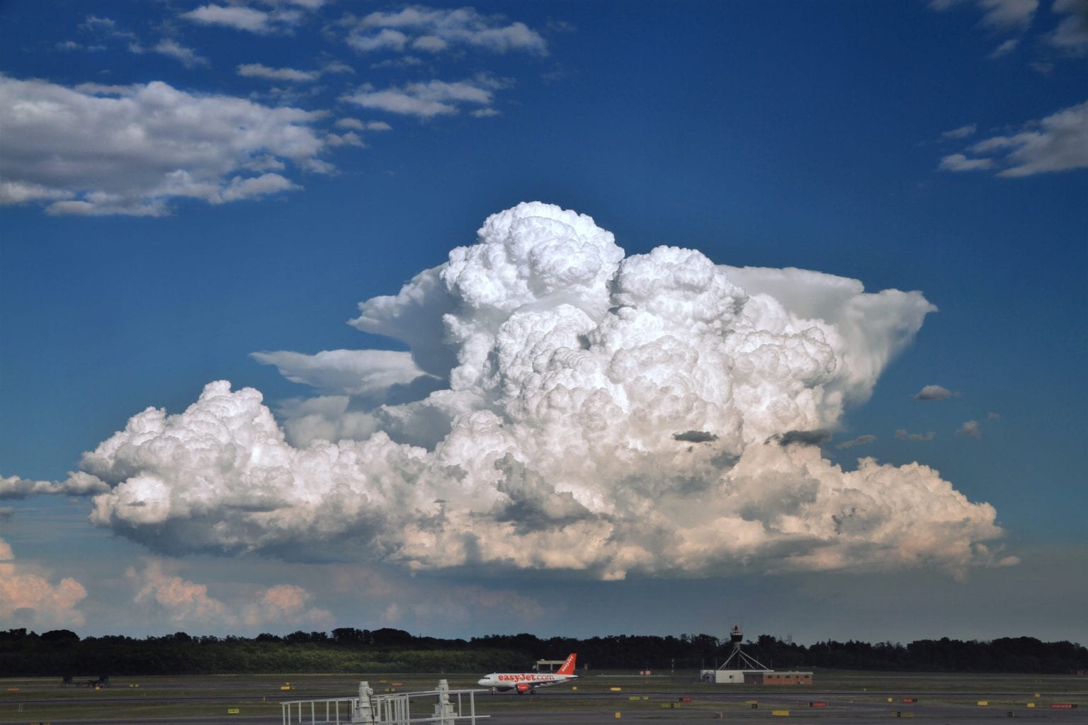 A small red airplane taxis on a runway under a large formation of white cumulus clouds against a blue sky.