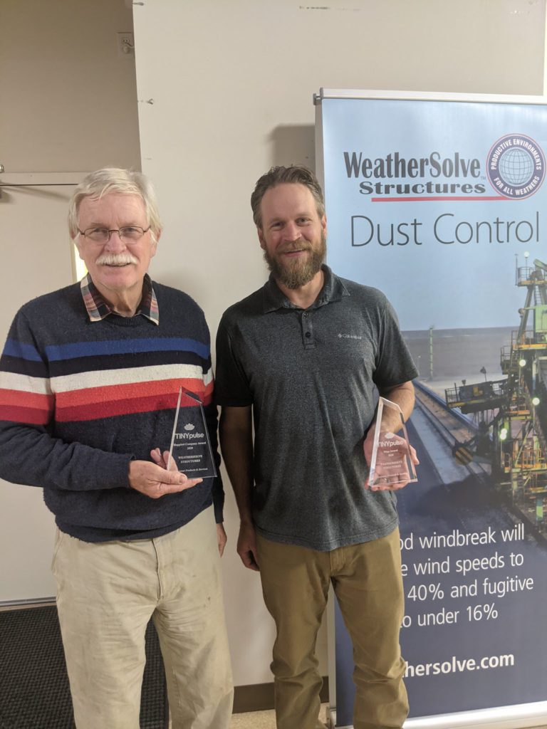 Two WeatherSolve team members holding TINYpulse awards in front of a WeatherSolve Structures Dust Control banner