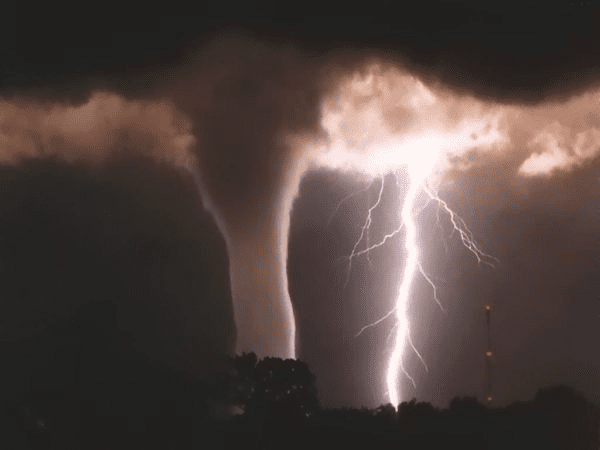 A tornado and lightning bolt occur simultaneously at night, with dark clouds above and silhouettes of trees below.