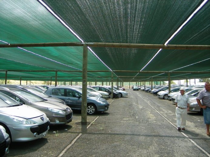 A parking lot with cars shaded by green fabric canopies supported by wooden poles