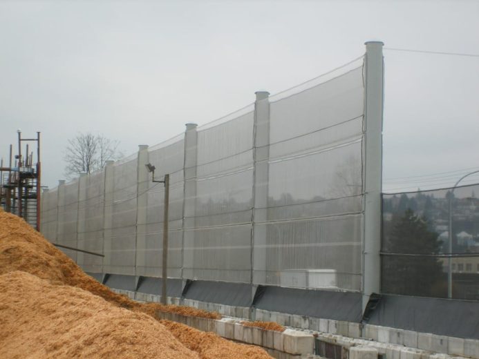 A tall, translucent wind fence stretches along a construction site with a pile of wood chips in the foreground