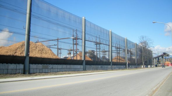 A long industrial fence runs alongside a road, with piles of material behind it