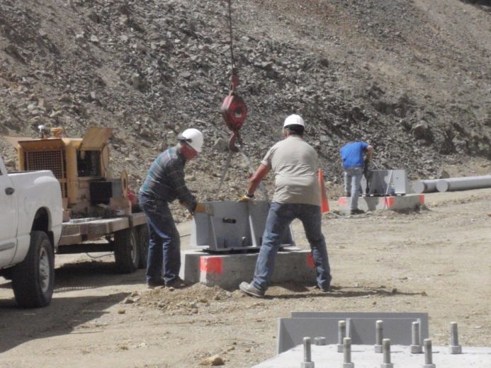 Workers in hard hats handle large metal components at a dusty construction site