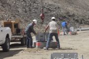 Workers in hard hats handle large metal components at a dusty construction site