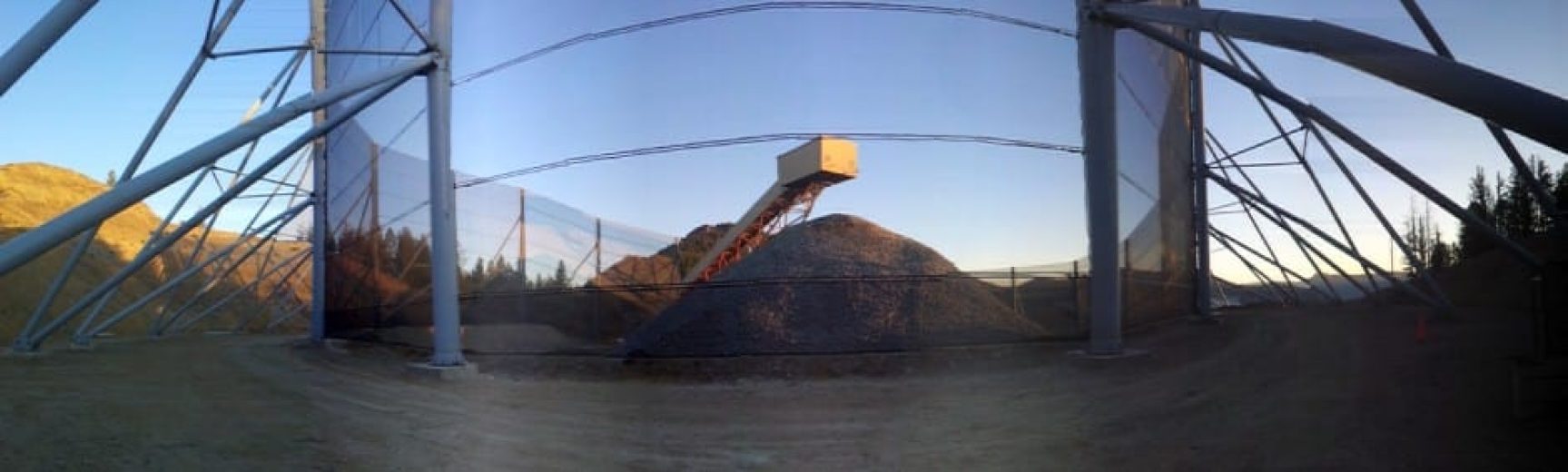 A large pile of gravel is covered by a netted structure supported by metal poles
