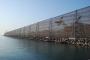 A long, transparent fence runs along a rocky shoreline with a crane visible behind it
