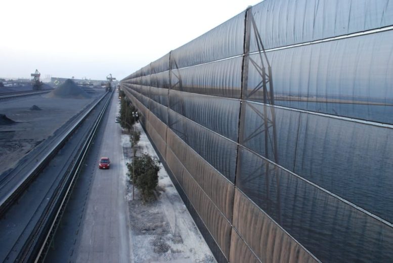 A long industrial wind fence stretches alongside a road with a red car driving past
