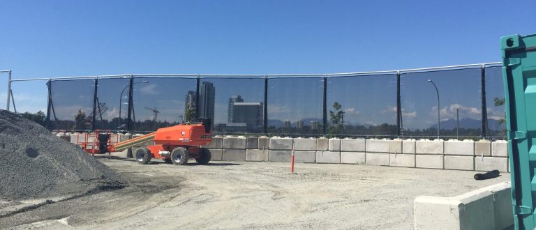 Construction site with orange machinery, gravel, concrete barriers, and dark mesh fencing