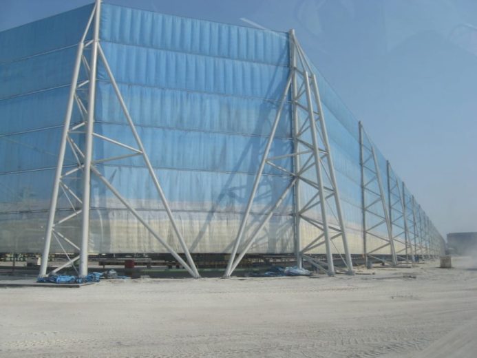 A large blue industrial wind fence supported by white metal framework stands on a dusty ground