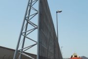 A tall, triangular metal structure stands next to a road with an orange JLG lift nearby