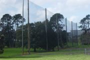 Tall poles with netting stretch across a grassy area with trees and a building in the background
