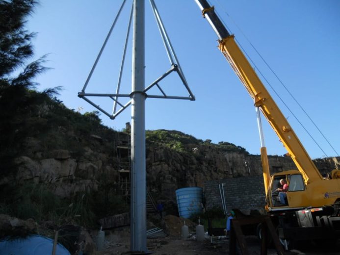 A crane lifts a tall metal pole with triangular supports against a clear blue sky