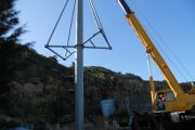 A crane lifts a tall metal pole with triangular supports against a clear blue sky