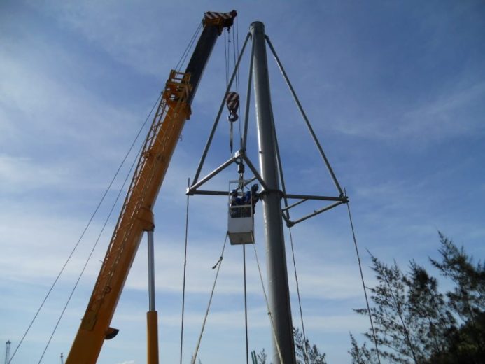 A crane lifts workers in a basket to the top of a tall metal pole