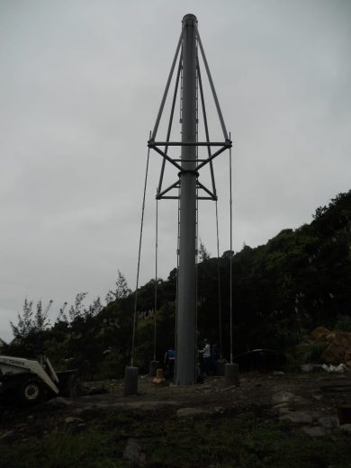 A tall metal pole with guy wires and a ladder, surrounded by construction equipment and workers