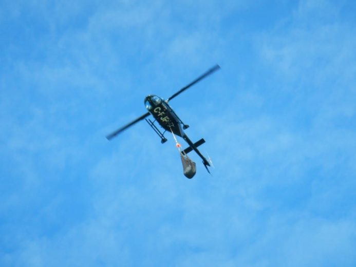 A helicopter flies against a blue sky with a bucket suspended below it