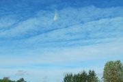A drone flies in a clear blue sky above a grassy area with trees and a building