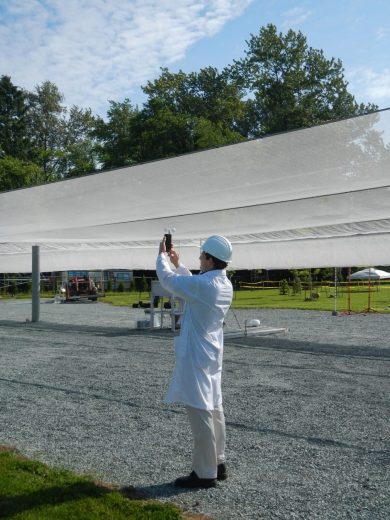 A person in a lab coat and hard hat takes a photo under a large white canopy