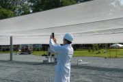 A person in a lab coat and hard hat takes a photo under a large white canopy