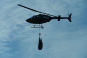 A helicopter lifts a large, dark object suspended by cables against a cloudy sky
