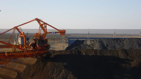 A large industrial machine operates near a massive pile of dark material, possibly coal or ore, under a clear sky