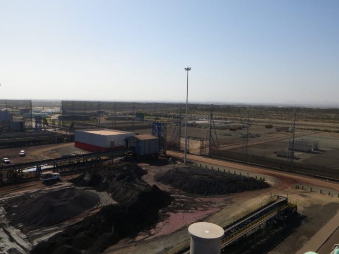 An industrial site with piles of dark material, buildings, and fencing under a clear sky
