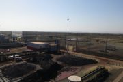 An industrial site with piles of dark material, buildings, and fencing under a clear sky