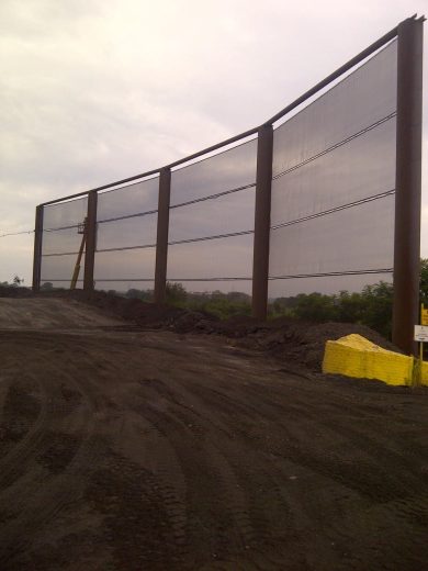 A large metal fence structure stands on a dirt ground with a cloudy sky in the background