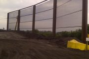 A large metal fence structure stands on a dirt ground with a cloudy sky in the background