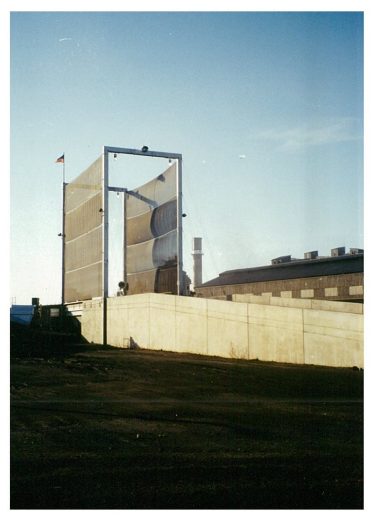 A large industrial wind fence stands against a clear sky, with a concrete wall and industrial buildings in the background