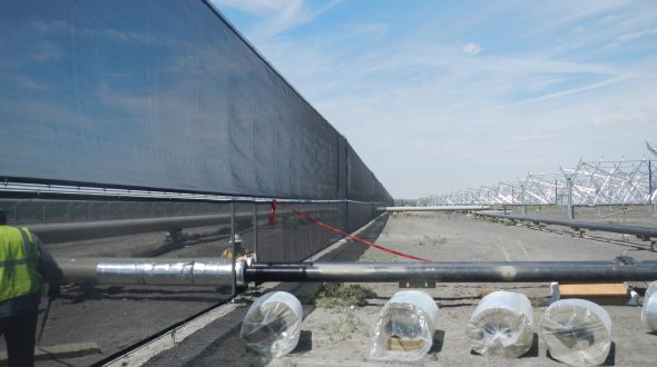 A long black wind fence stretches across a construction site with pipes and wrapped materials in the foreground