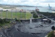 A coastal industrial area with a large wind fence, conveyor belt, and shipping cranes