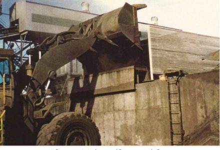 A large industrial loader pours material into a hopper with a wind fence and fog system in place