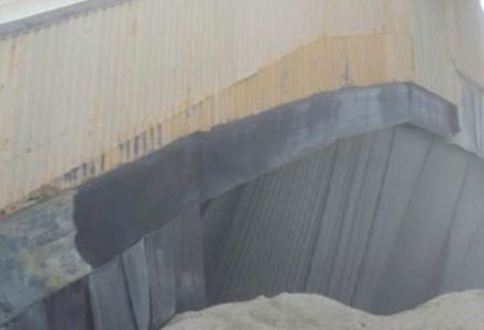 Corrugated metal siding with peeling paint and a dark patch, above a pile of light-colored material