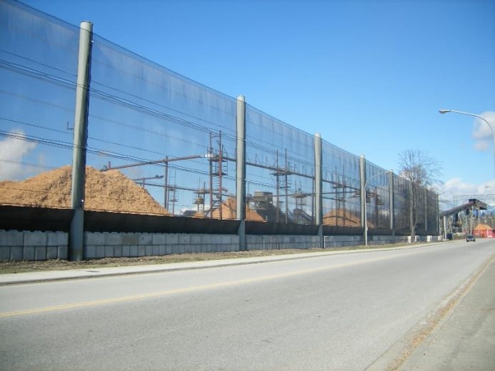 A long, tall wind fence stretches alongside a road, with piles of wood fiber visible behind it