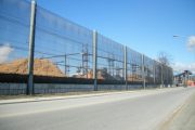 A long, tall wind fence stretches alongside a road, with piles of wood fiber visible behind it