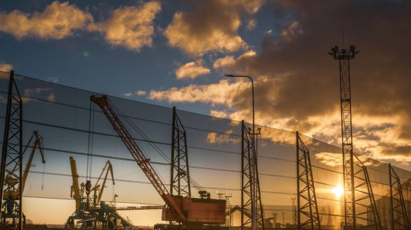 A construction site with cranes and a large wind fence at sunset