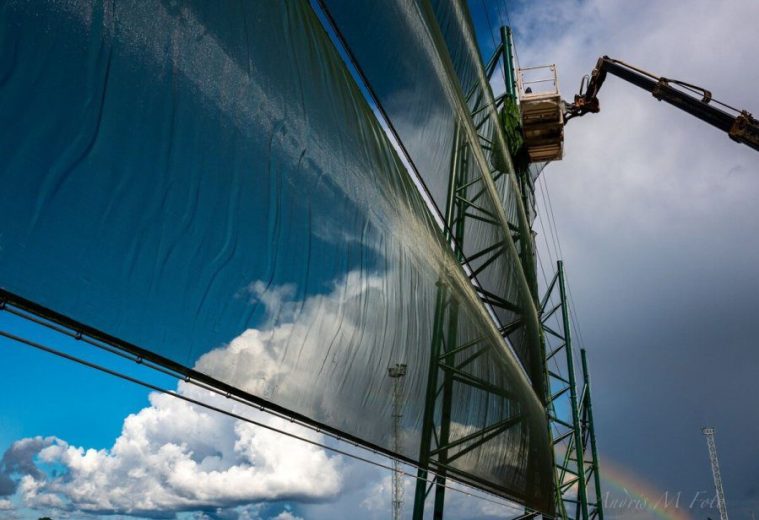A large green metal structure with a translucent covering against a backdrop of clouds and a rainbow
