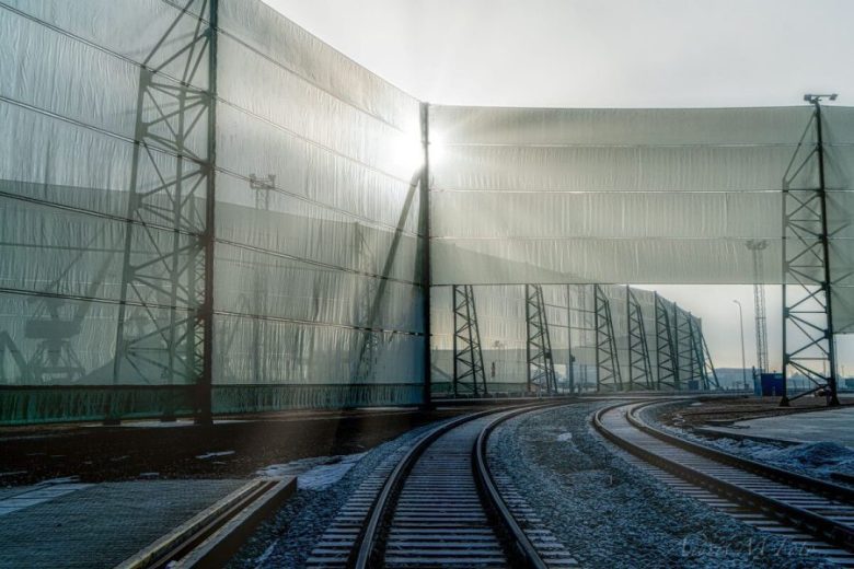 Railway tracks curve gently beside tall, translucent wind fences, illuminated by sunlight